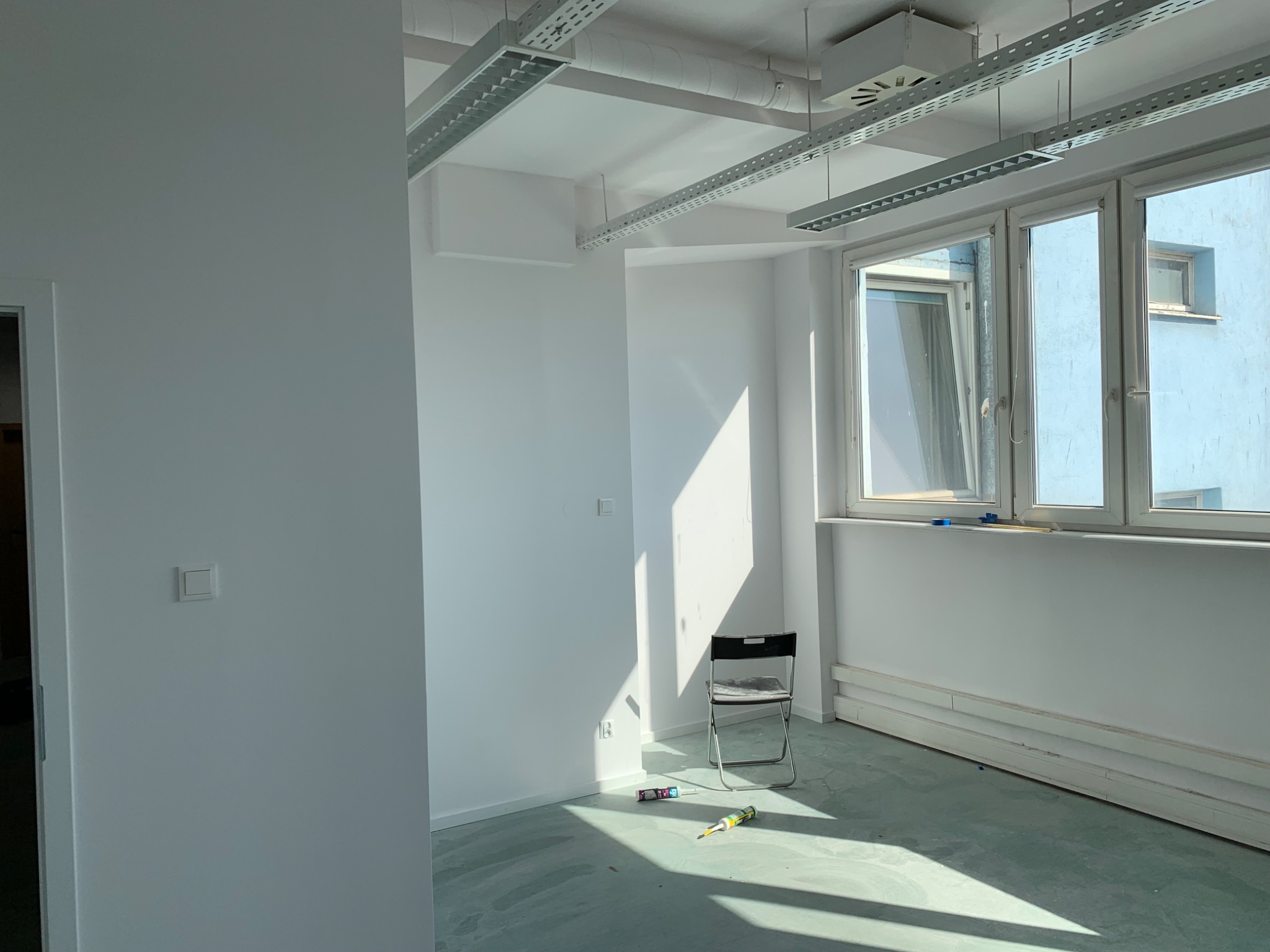 Puste, jasne pomieszczenie w trakcie przygotowań do prac wykończeniowych; widoczne listwy przypodłogowe, krzesło składane, tuba z klejem na podłodze i mocne promienie słoneczne wpadające przez okno.