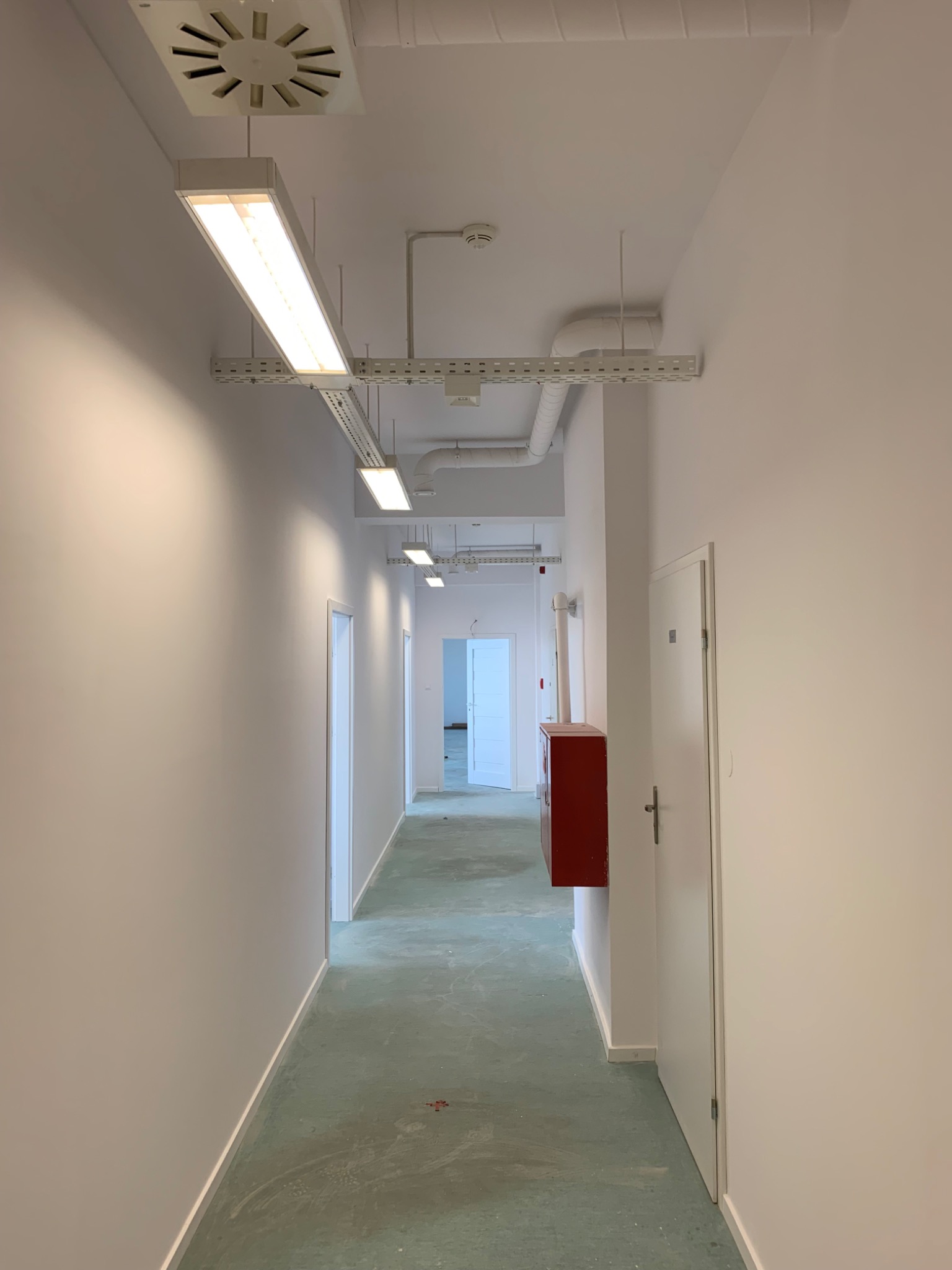 Długi, jasny korytarz z białymi ścianami, podwieszanymi lampami i zieloną podłogą, widoczna instalacja elektryczna pod sufitem oraz czerwona szafka hydrantowa przy ścianie.