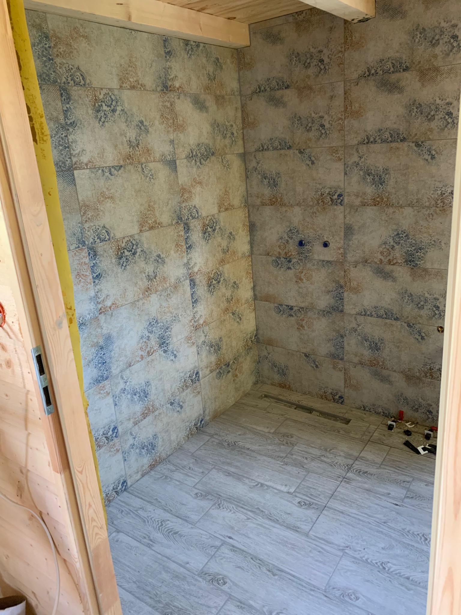 Niedokończona łazienka z ułożonymi płytkami ściennymi w kwiatowy wzór i podłogowymi imitującymi drewno, widoczne elementy instalacji hydraulicznej i narzędzia glazurnicze.