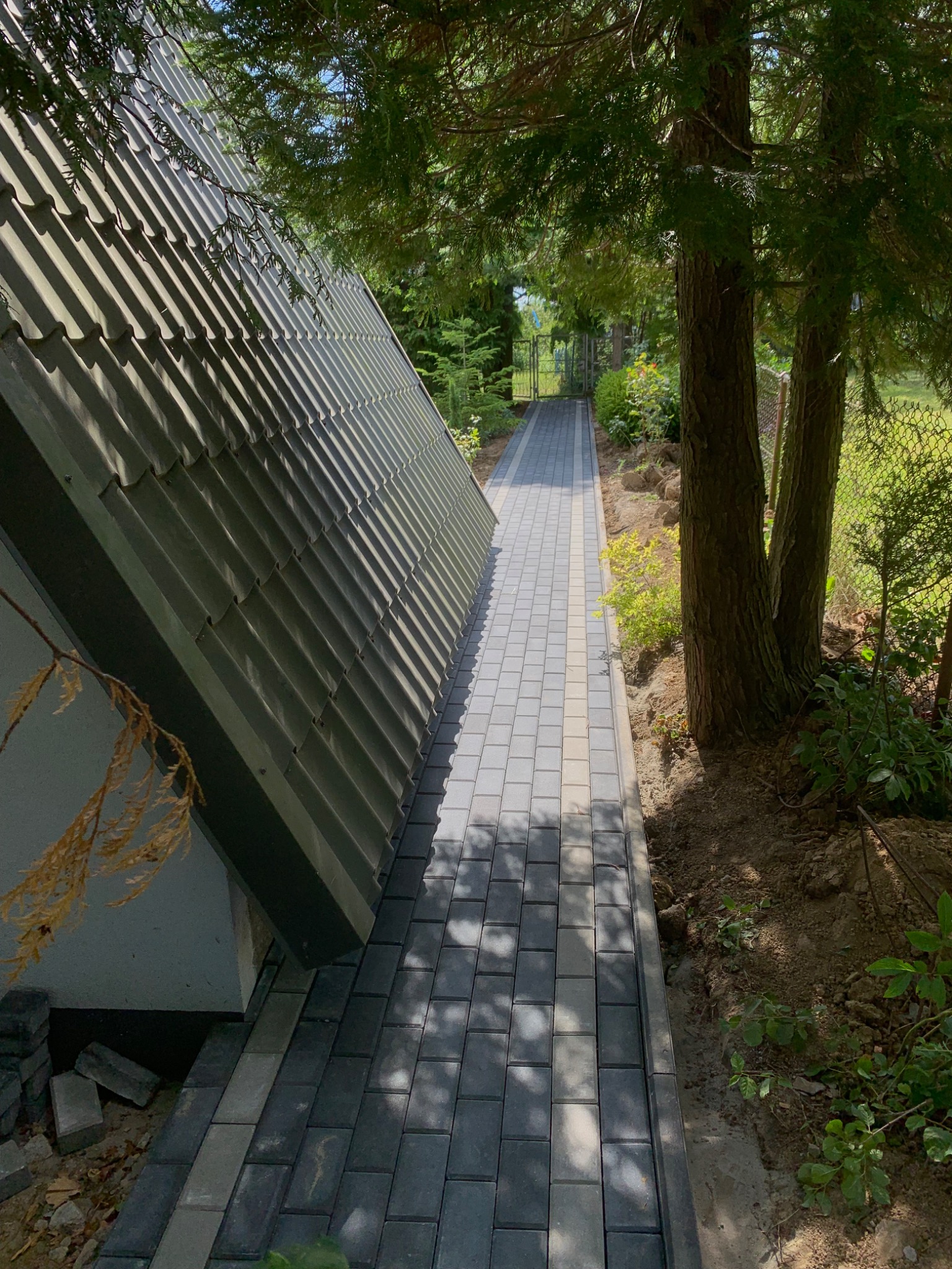 Ścieżka z szarych prostokątnych kostek brukowych, z jasnym pasem pośrodku, prowadząca wzdłuż ściany budynku z ciemnym, spadzistym dachem, otoczona zielenią i drzewami.