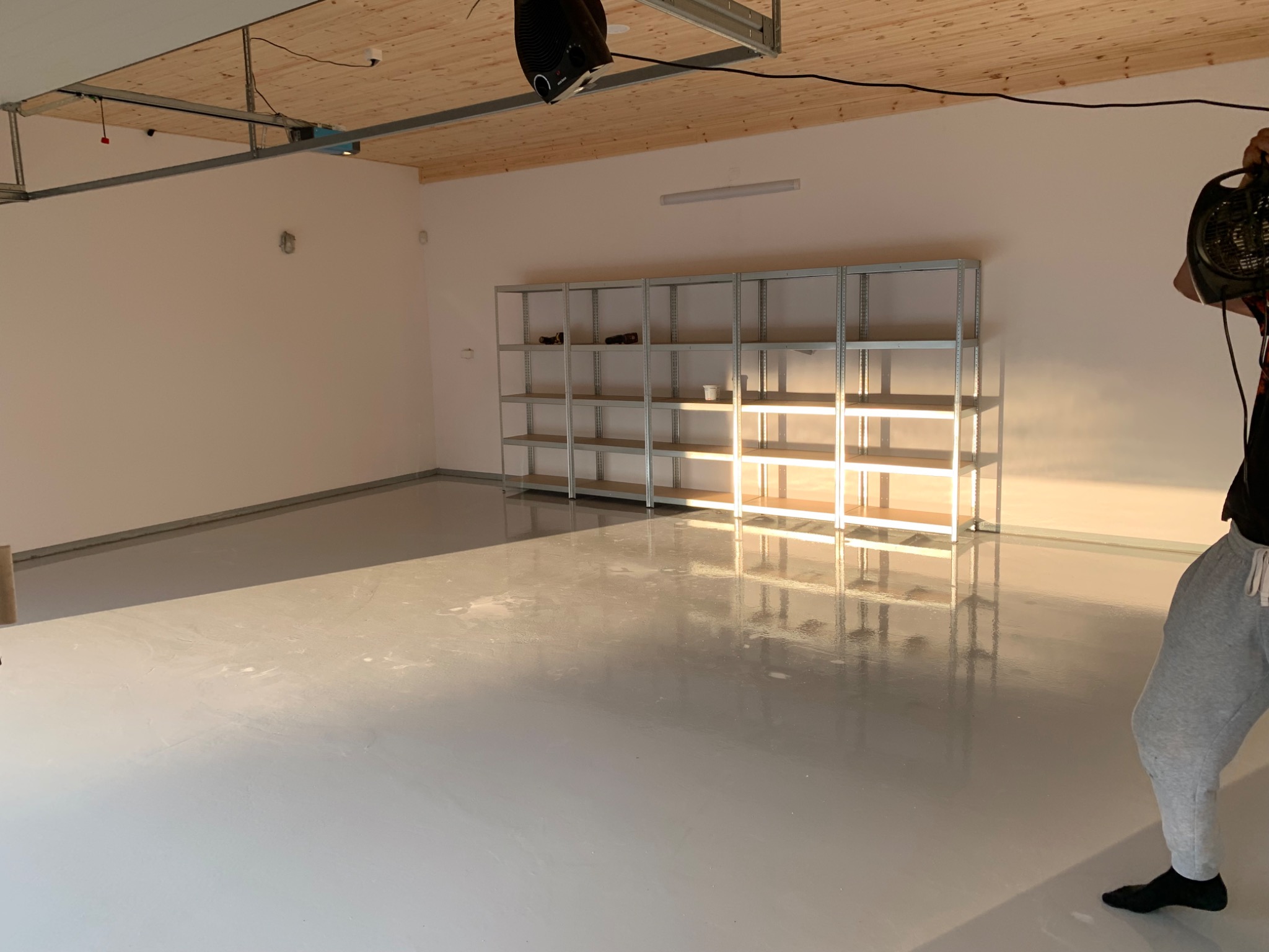 Świeżo wylana, błyszcząca, szara posadzka epoksydowa w pustym pomieszczeniu z metalowymi regałami, z widocznym fragmentem osoby trzymającej nagrzewnicę.