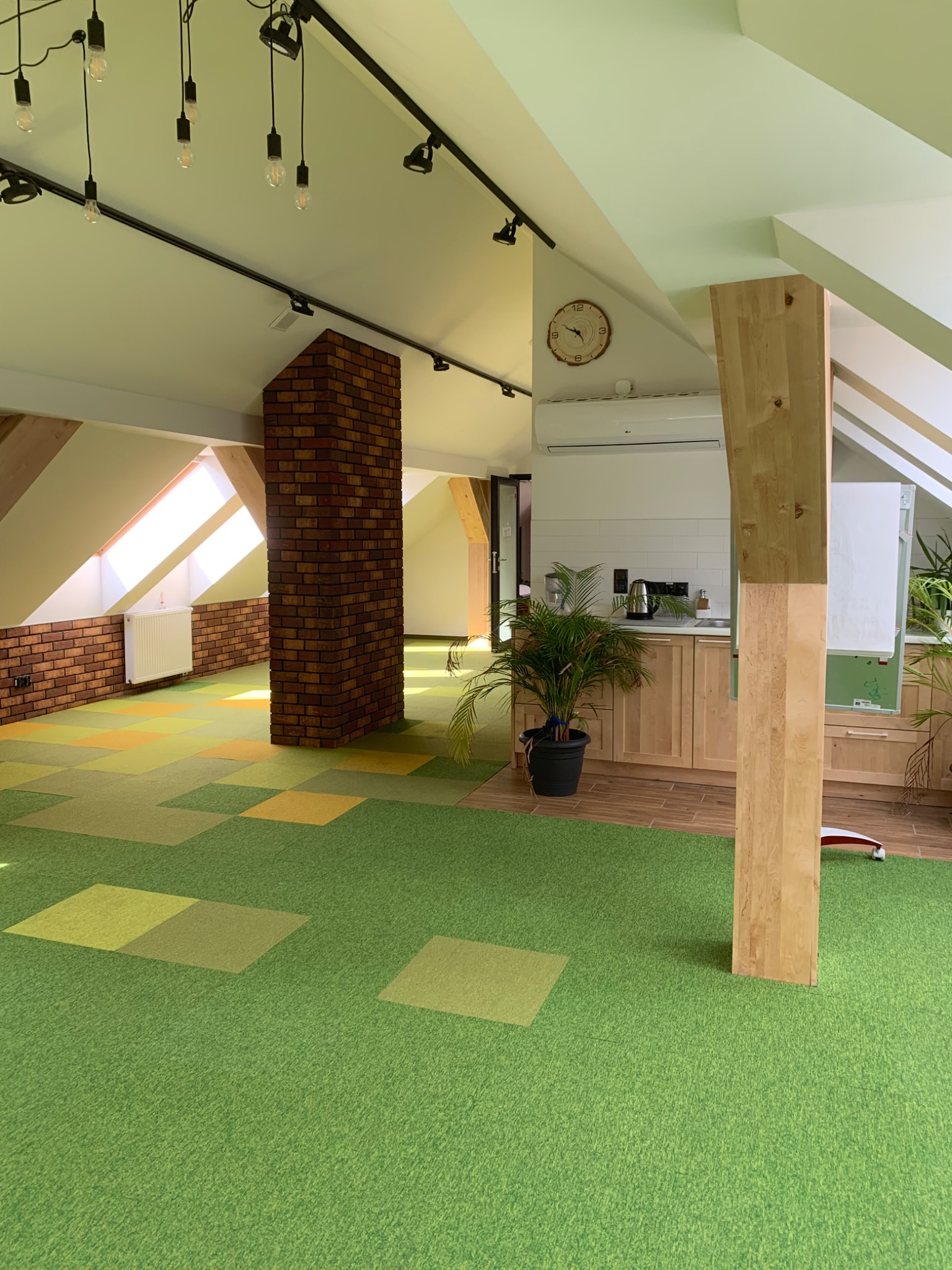 Przestronne poddasze w Tczewie po remoncie, z zieloną wykładziną dywanową w kwadraty, ceglanym filarem i ścianą, drewnianymi belkami oraz aneksem kuchennym w tle.