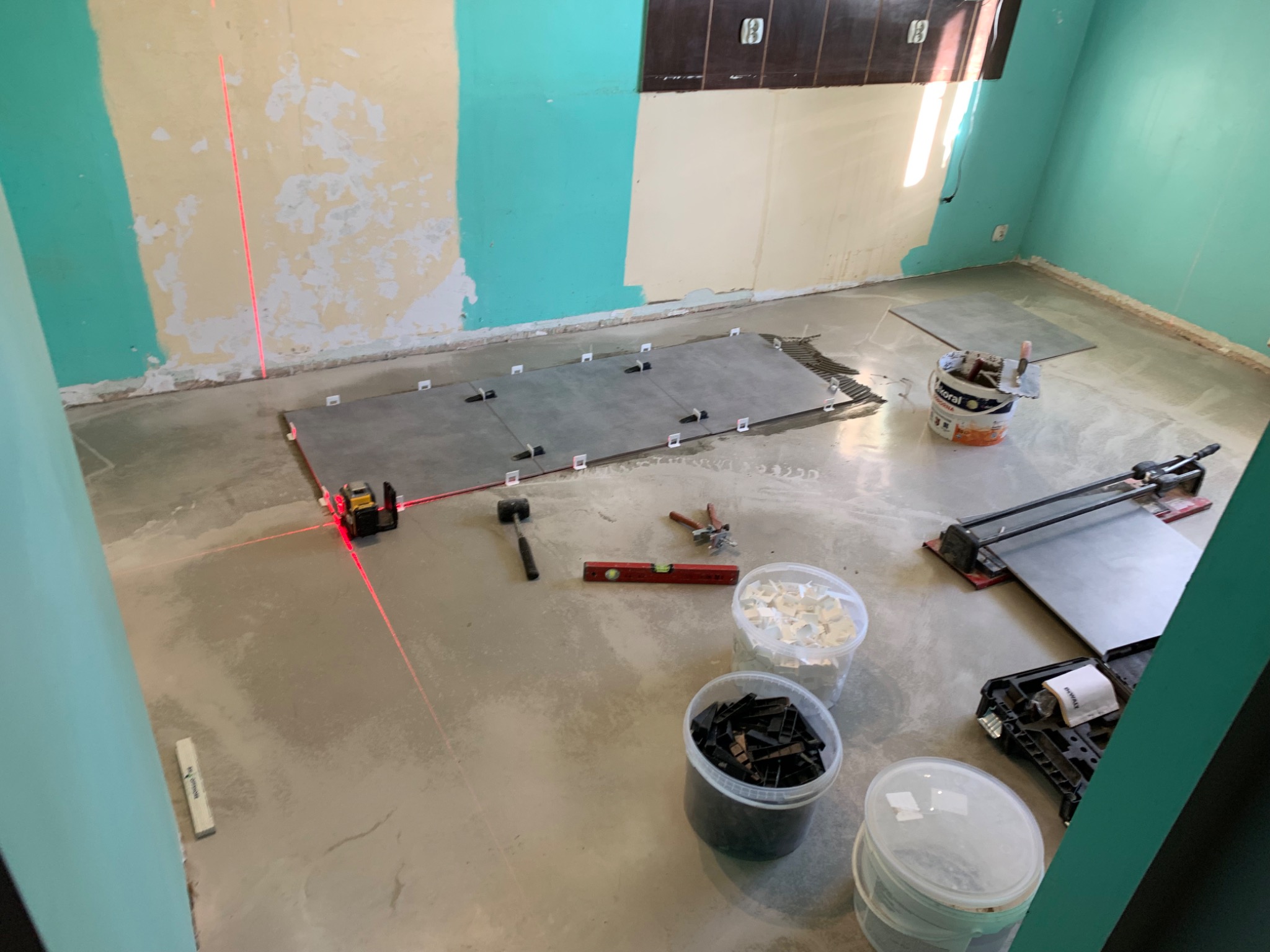 Układanie szarych płytek podłogowych w pomieszczeniu z pomalowanymi na biało-turkusowo ścianami, widoczny laser krzyżowy wyznaczający poziom, poziomica, młotek gumowy, wiaderka z krzyżykami...