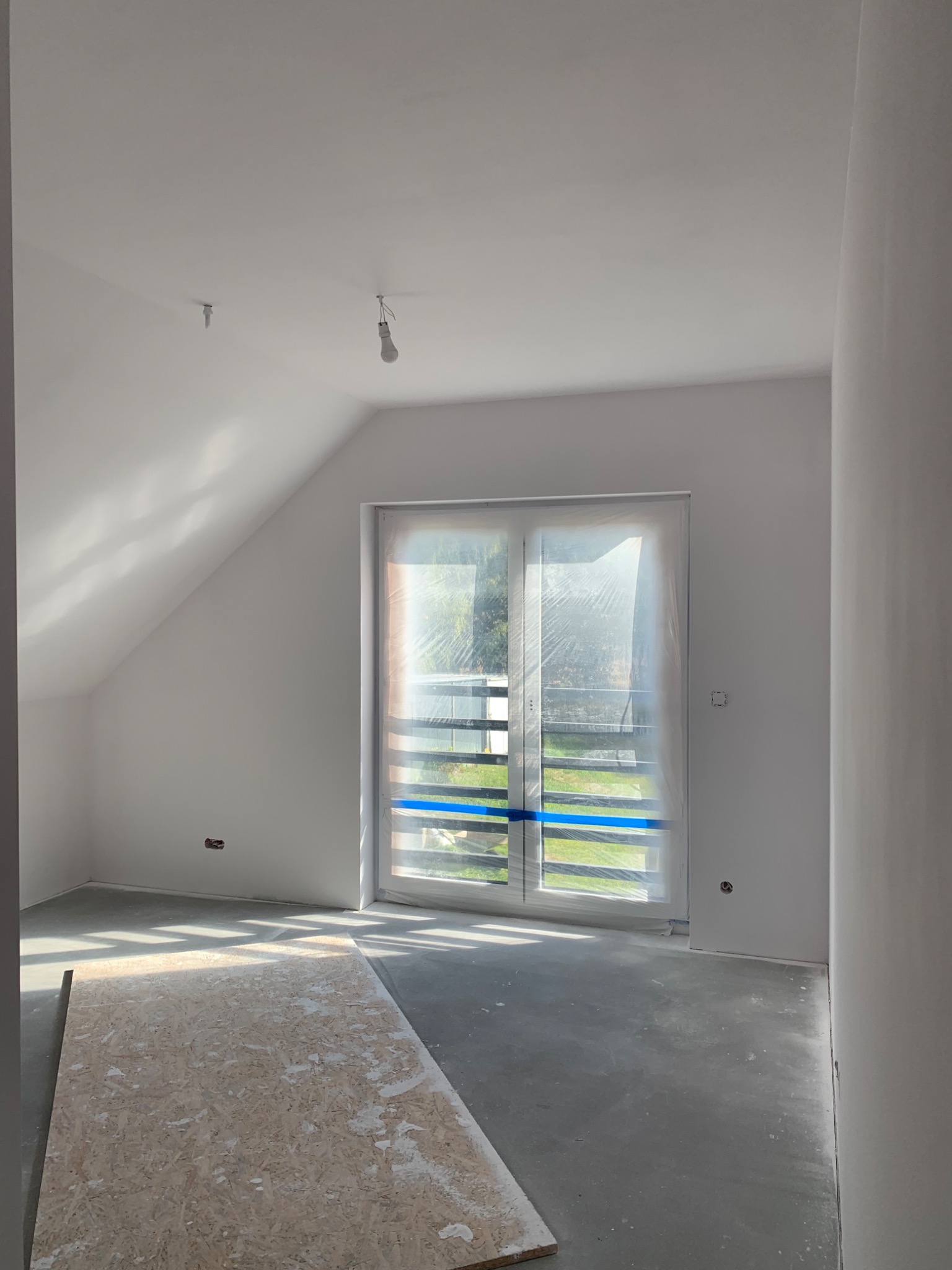 Pokój na poddaszu w trakcie prac wykończeniowych; okno zabezpieczone folią, ściany świeżo pomalowane na biało, widoczne gniazdka elektryczne bez osprzętu, na podłodze płyta OSB.