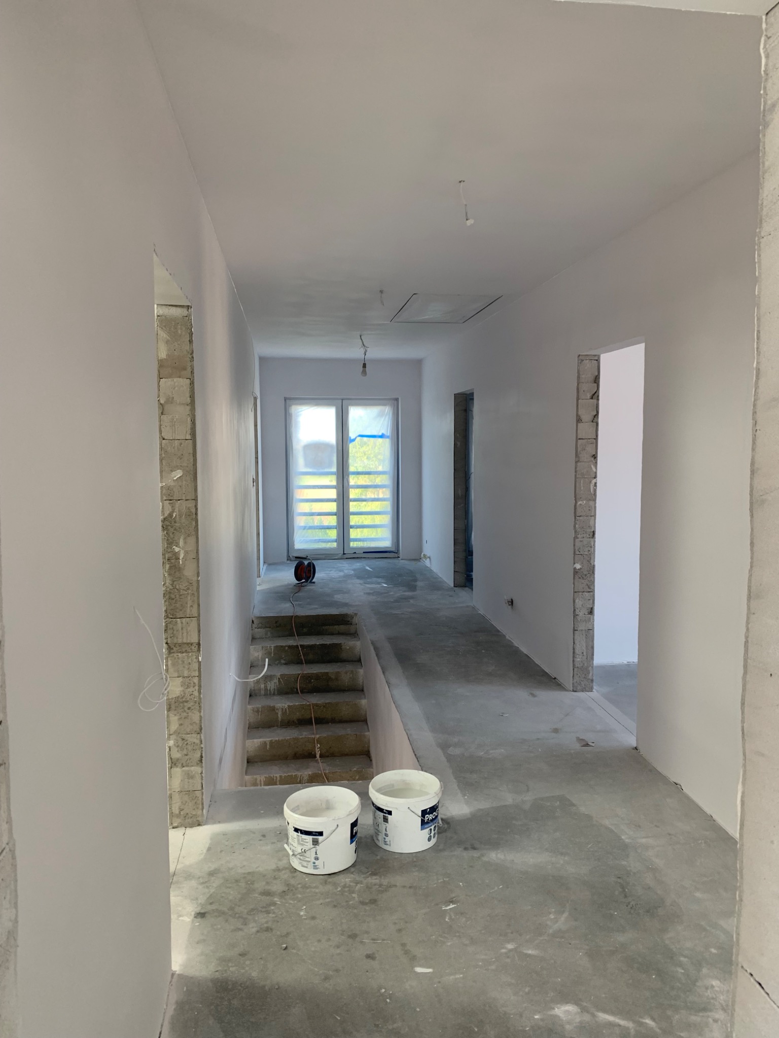 Korytarz w trakcie remontu: ściany po szpachlowaniu i malowaniu na biało, widoczne dwa wiadra z farbą, fragment schodów z zabezpieczonym oknem w tle, surowy beton na podłodze.