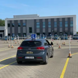 Szary samochód Kia Rio z niebieską literą L na dachu, zaparkowany na placu manewrowym z pomarańczowymi pachołkami i żółtymi liniami, w tle budynek szkoły.