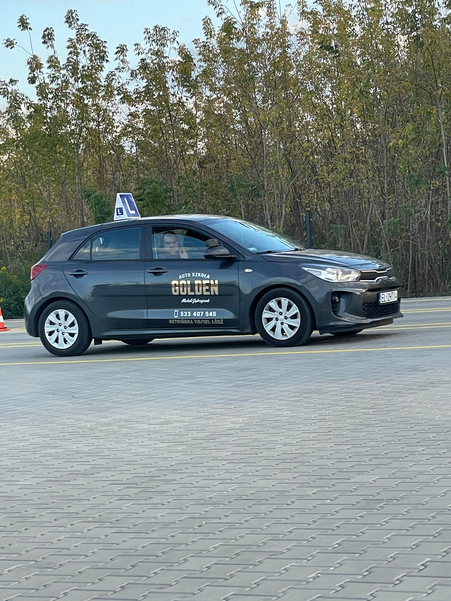 Szary samochód z oznaczeniami szkoły jazdy Golden z Łodzi, z charakterystycznym znakiem 'L' na dachu, zaparkowany na brukowanej powierzchni z zielonymi drzewami w tle.