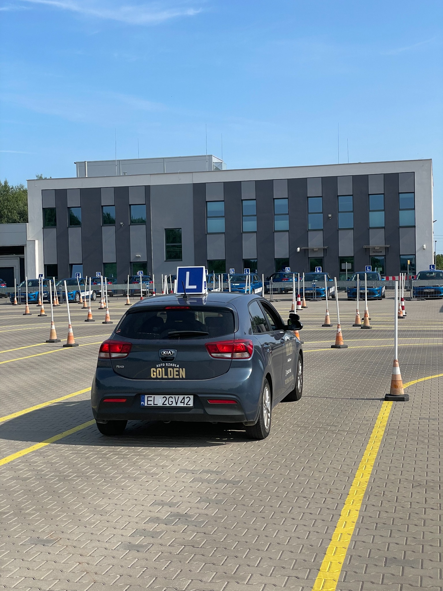 Szary samochód Kia Rio z niebieską literą L na dachu, zaparkowany na placu manewrowym z pomarańczowymi pachołkami i żółtymi liniami, w tle budynek szkoły.