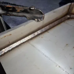 Detal z procesu spawania metalowej konstrukcji, widoczny świeży spaw o opalizującym zabarwieniu oraz uchwyt spawalniczy w górnej części kadru.