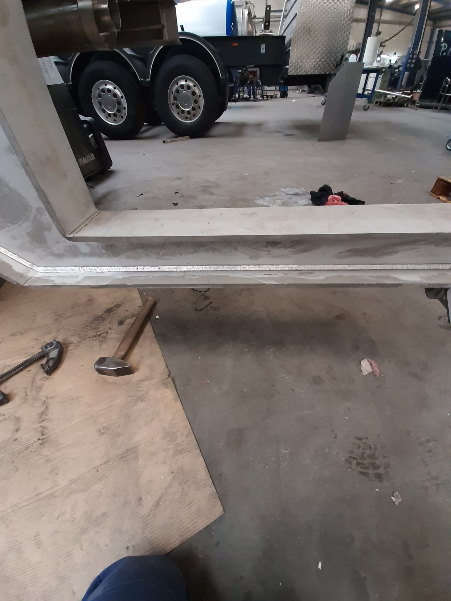 Fragment spawanej konstrukcji metalowej w warsztacie, widoczne narzędzia: młotek i klucz, w tle koła ciężarówki.