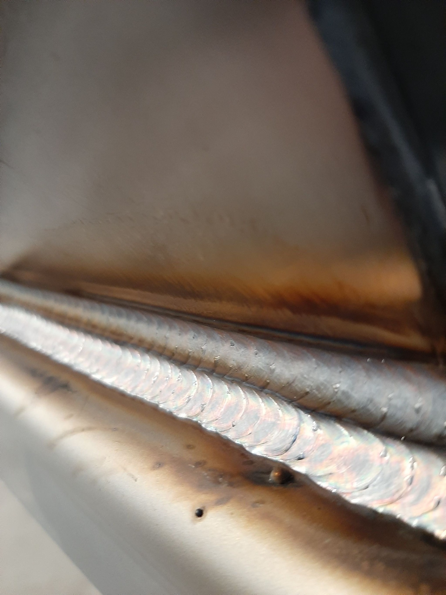 Zbliżenie spoiny na metalu, widoczne charakterystyczne ślady po spawaniu w formie nakładających się półksiężyców, z lekkim odbarwieniem termicznym metalu.