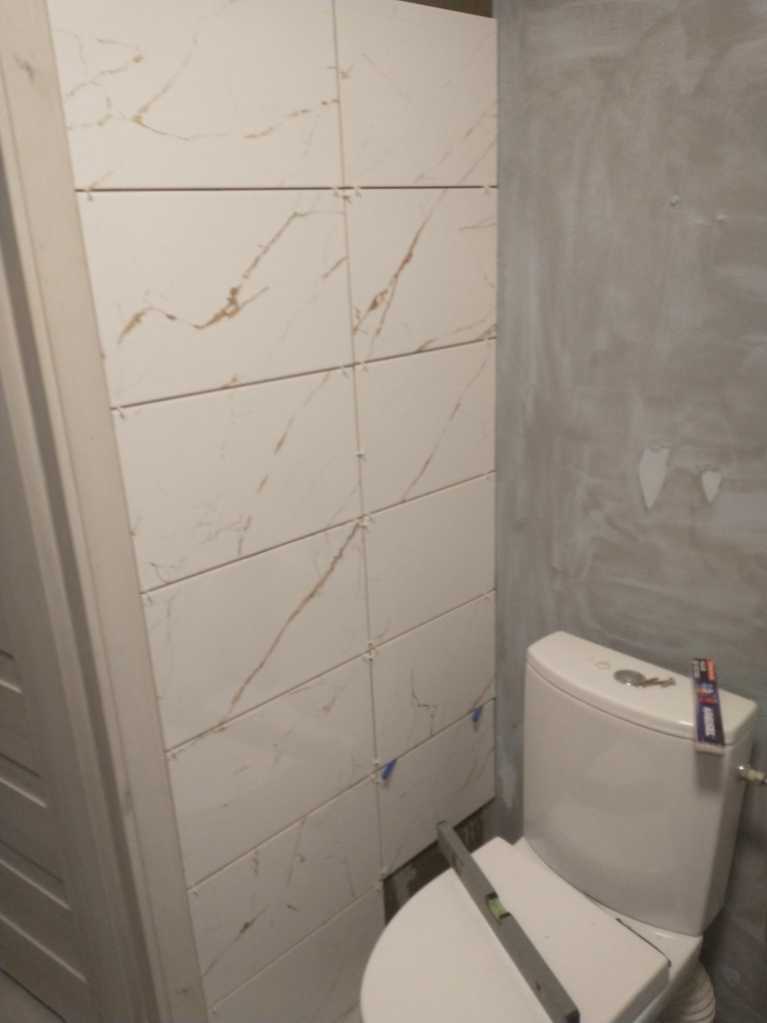 Świeżo ułożone, beżowe płytki imitujące marmur na ścianie w łazience, z niezagruntowaną ścianą obok i poziomnicą na sedesie.