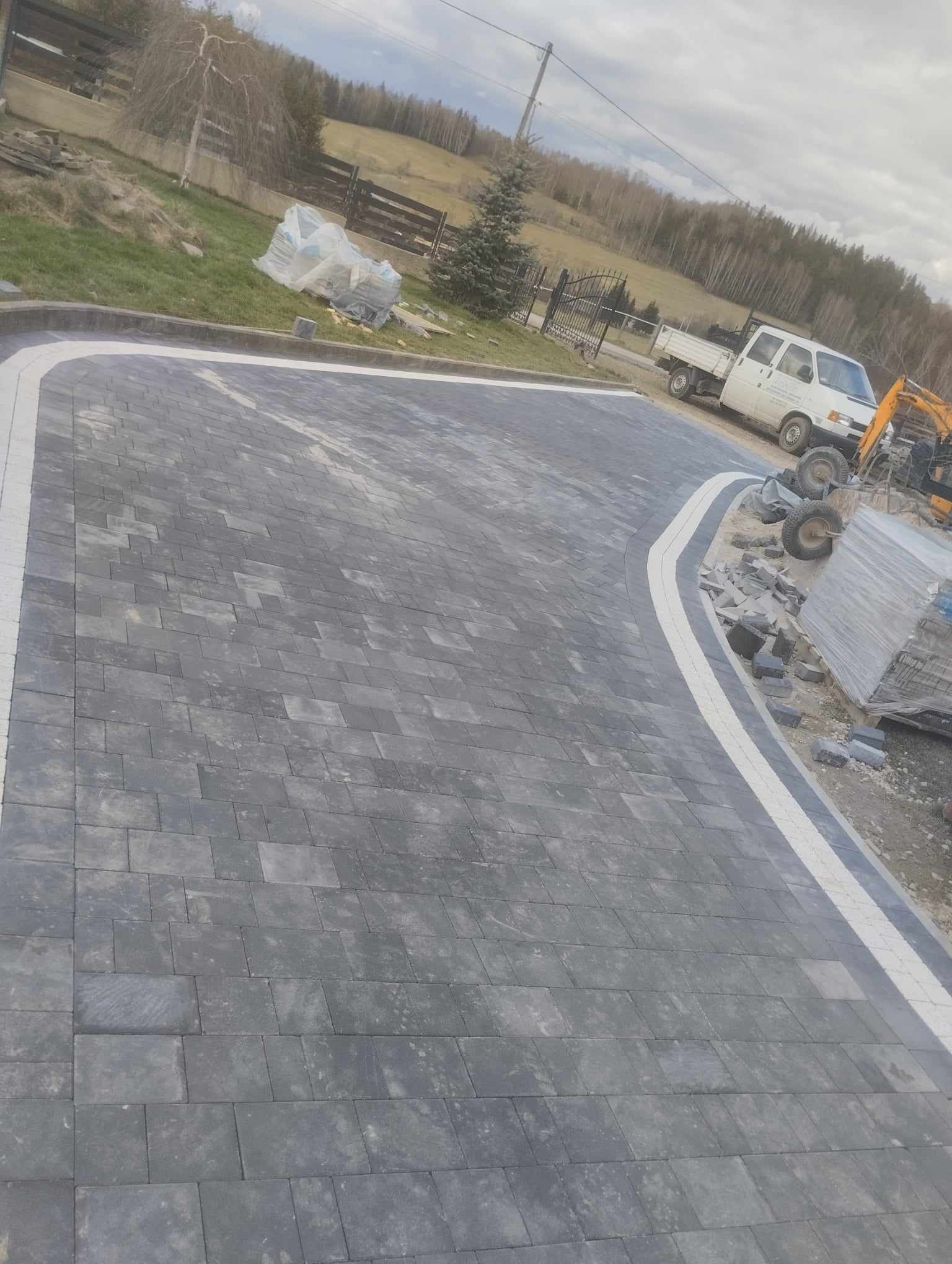 Ukończona nawierzchnia z szarej kostki brukowej z białym obramowaniem, widoczny samochód dostawczy i mini koparka w tle, teren lekko pofałdowany.