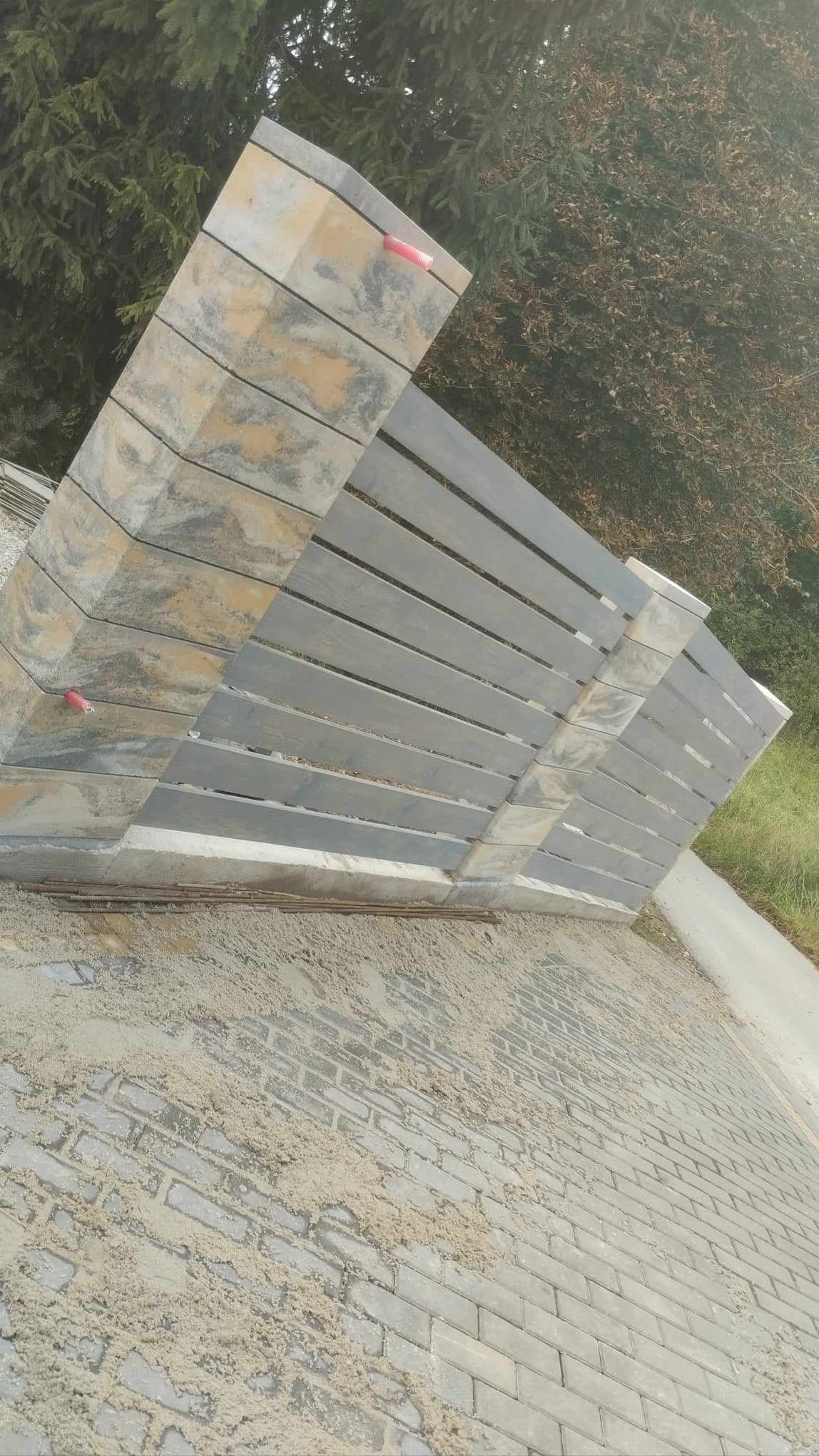Ułożona kostka brukowa z szarymi, poziomymi elementami ogrodzenia w tle, widoczne ślady piasku na kostce i czerwony znacznik na elemencie ogrodzenia.