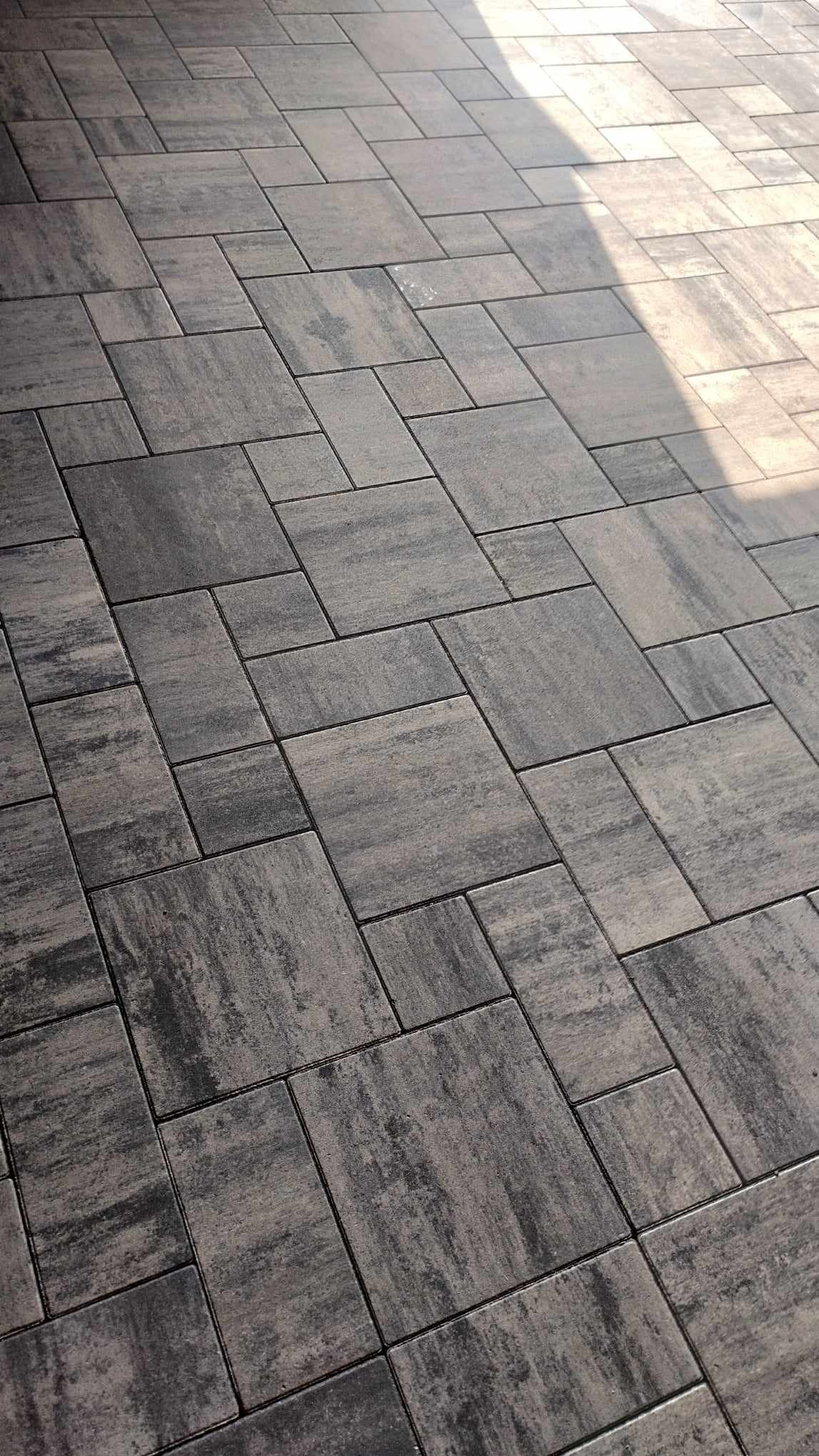 Ułożona nawierzchnia z prostokątnych, szarych kostek brukowych o zróżnicowanej fakturze i wzorze, częściowo oświetlona przez słońce padające z góry.
