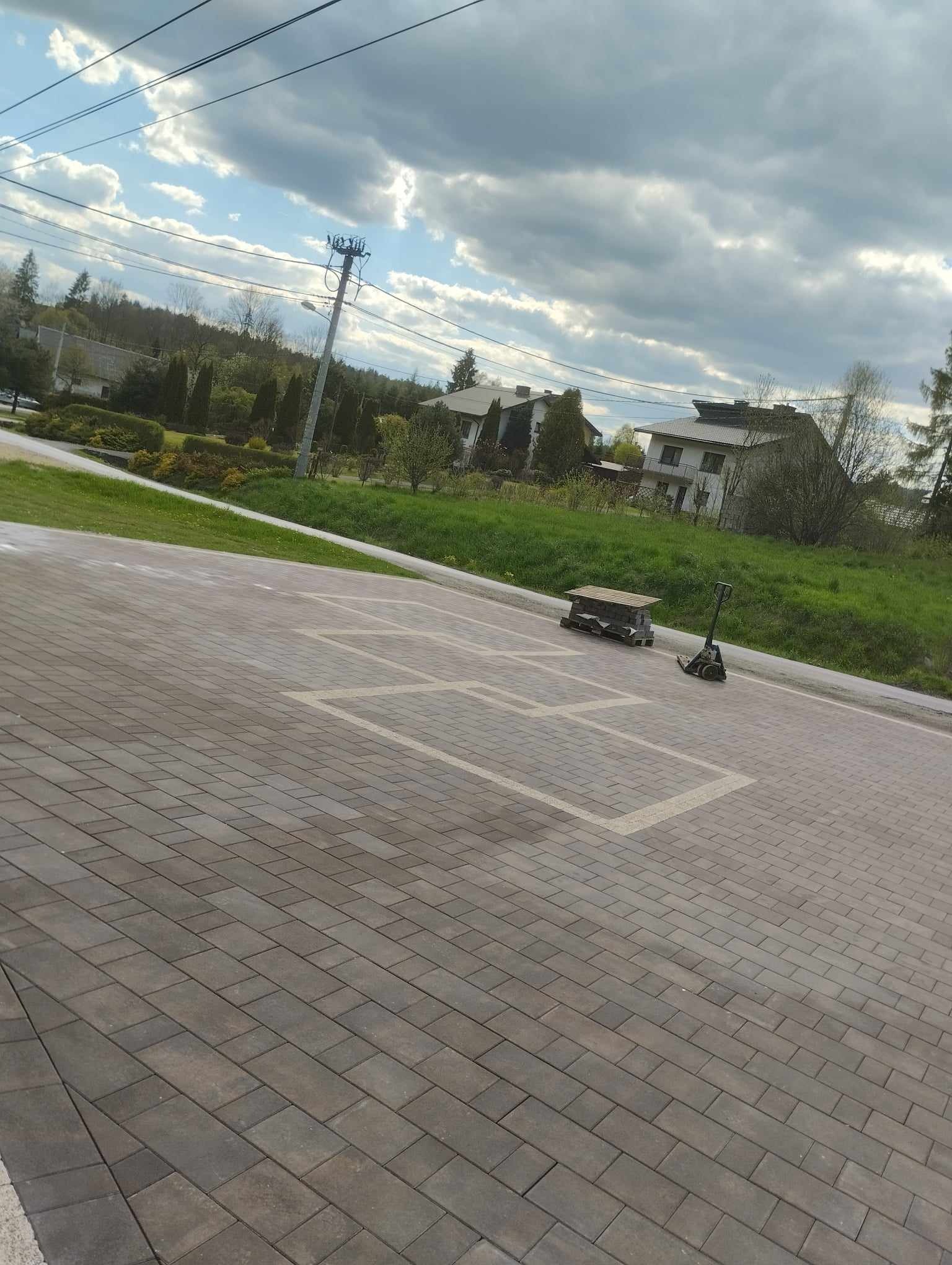 Ułożona kostka brukowa w odcieniach szarości i brązu, z zaznaczonymi białymi liniami polami, paleta z kostką i wózkiem transportowym w tle, domy jednorodzinne i zieleń w tle, pochmurne niebo.