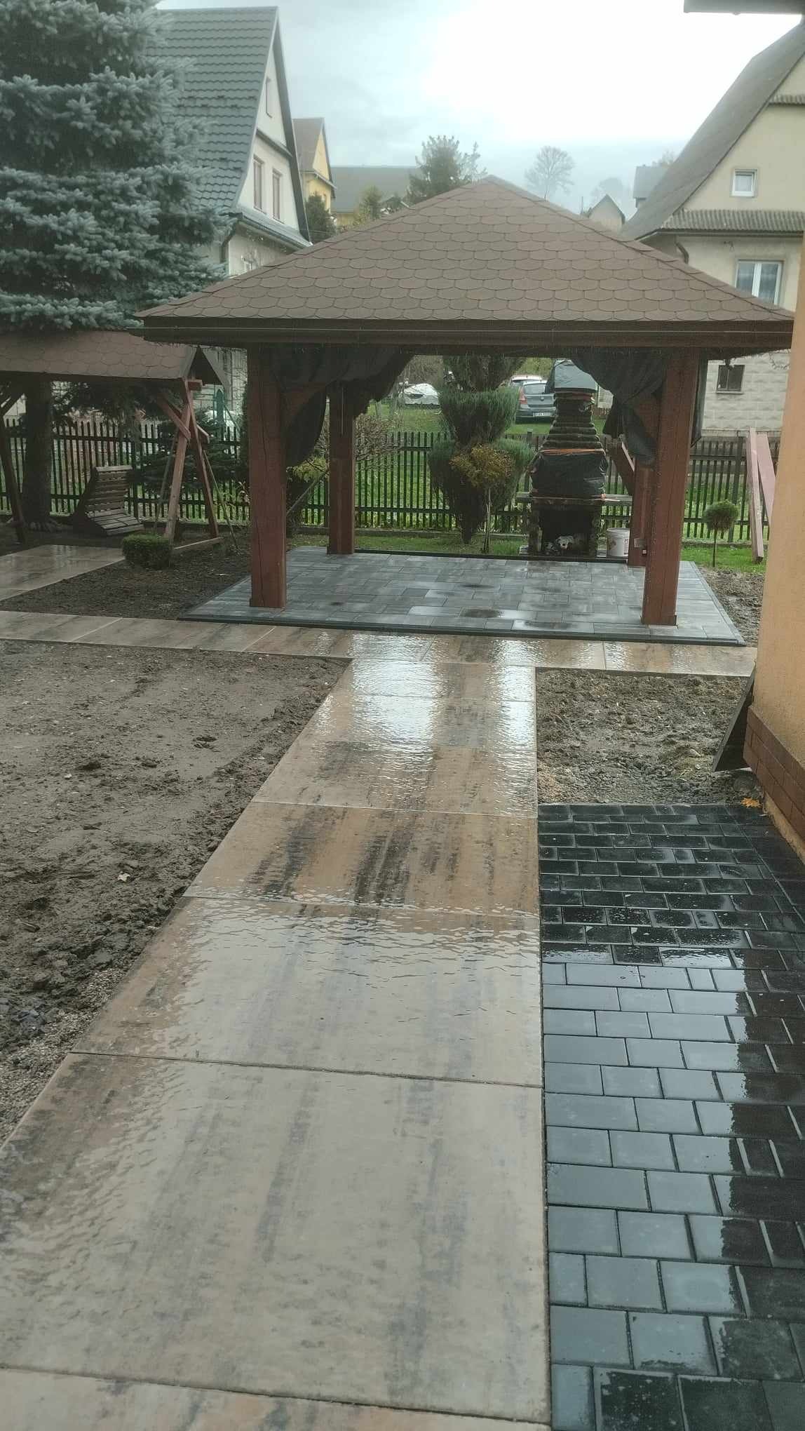 Świeżo ułożona, mokra kostka brukowa w dwóch odcieniach szarości prowadząca do drewnianej altany z zasłonami; widoczny fragment ogrodu i budynku w tle.