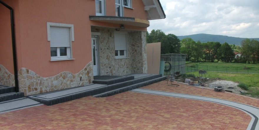 Ułożona kostka brukowa w odcieniach czerwieni i żółci przed domem z kamienną elewacją, widoczne schody wejściowe i fragment trawnika z widokiem na zalesione wzgórza w tle.