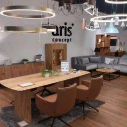 Wnętrze salonu meblowego z ekspozycją różnych modeli lamp wiszących, stołu z krzesłami oraz sofy narożnej w odcieniach szarości. Na ścianie logo firmy Aris Concept.