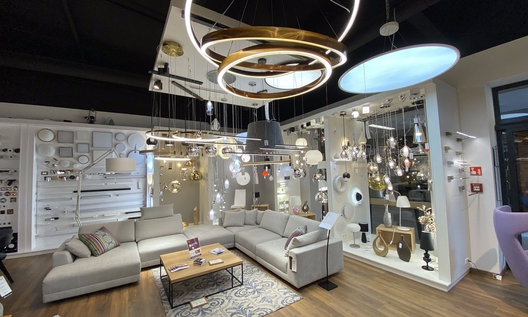 Salon ekspozycyjny z aranżacją różnorodnych lamp: wiszące, sufitowe, podłogowe i stołowe, w otoczeniu mebli wypoczynkowych.