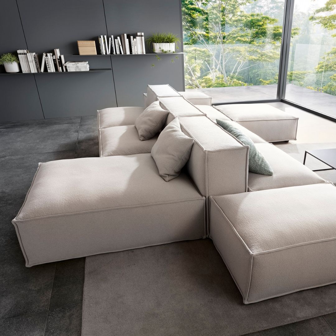 Modularna sofa w jasnych odcieniach z poduszkami, na tle dużego okna z widokiem na zieleń i półki z książkami.