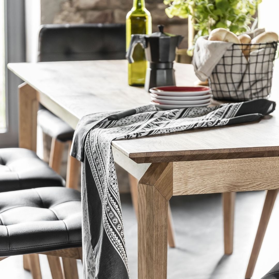 Drewniany stół z czarnymi skórzanymi krzesłami, nakryty wzorzystą szarą serwetą, w tle butelka oliwy, kawiarka i koszyk z pieczywem.