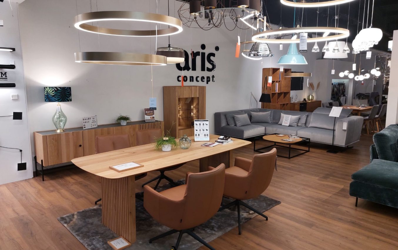 Wnętrze salonu meblowego z ekspozycją różnych modeli lamp wiszących, stołu z krzesłami oraz sofy narożnej w odcieniach szarości. Na ścianie logo firmy Aris Concept.