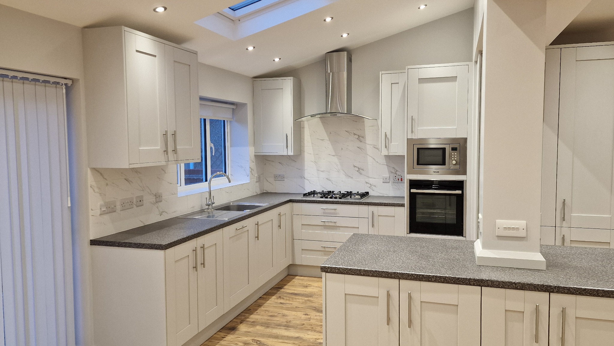 Jasna kuchnia w stylu skandynawskim z białymi szafkami, szarym blatem i panelami podłogowymi imitującymi drewno, okap ze stali nierdzewnej i marmurowe płytki na ścianie.