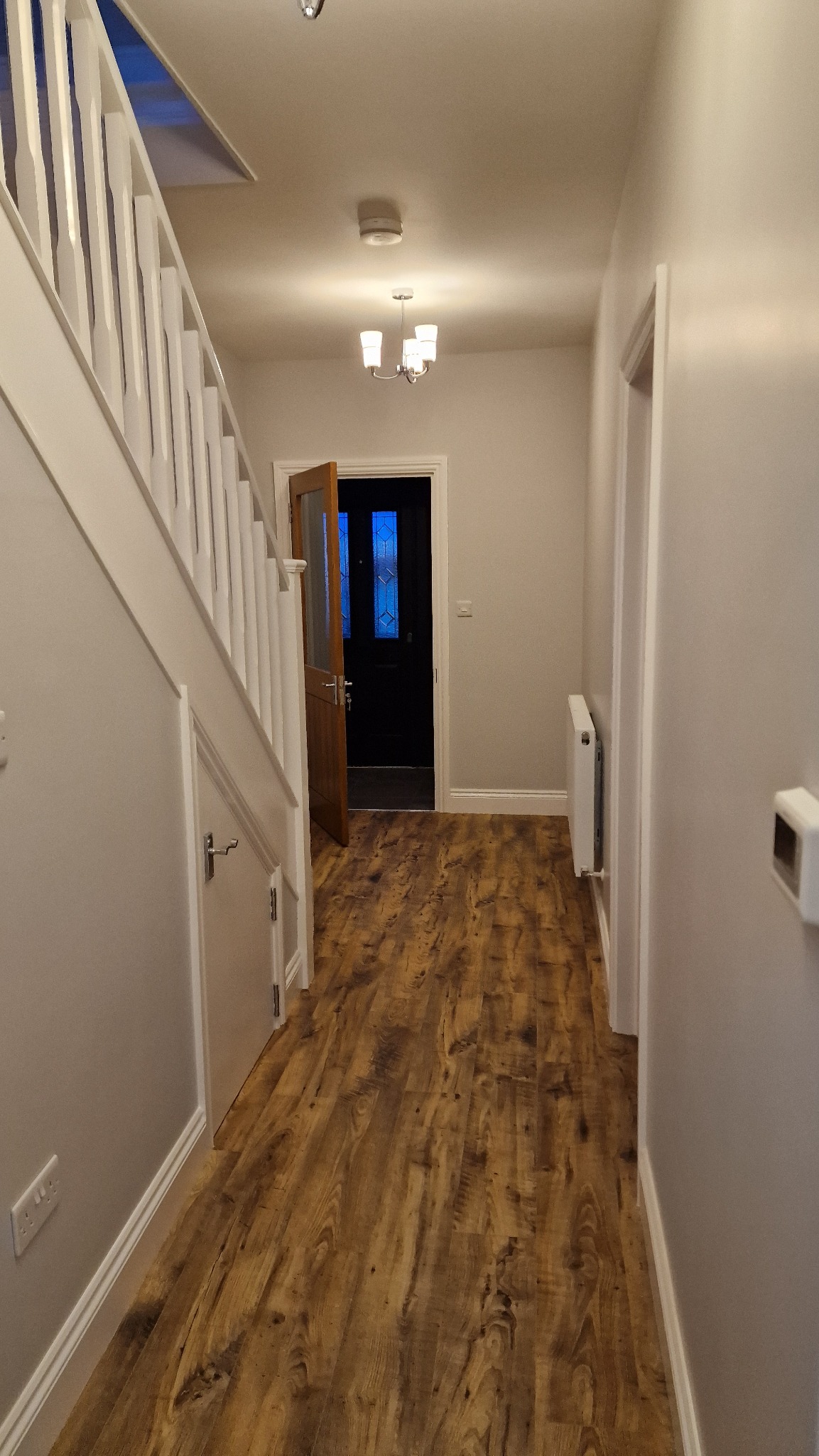 Wnętrze wyremontowanego korytarza z drewnopodobnymi panelami podłogowymi, białymi ścianami i schodami, widoczny grzejnik i drzwi wejściowe z przeszkleniami.