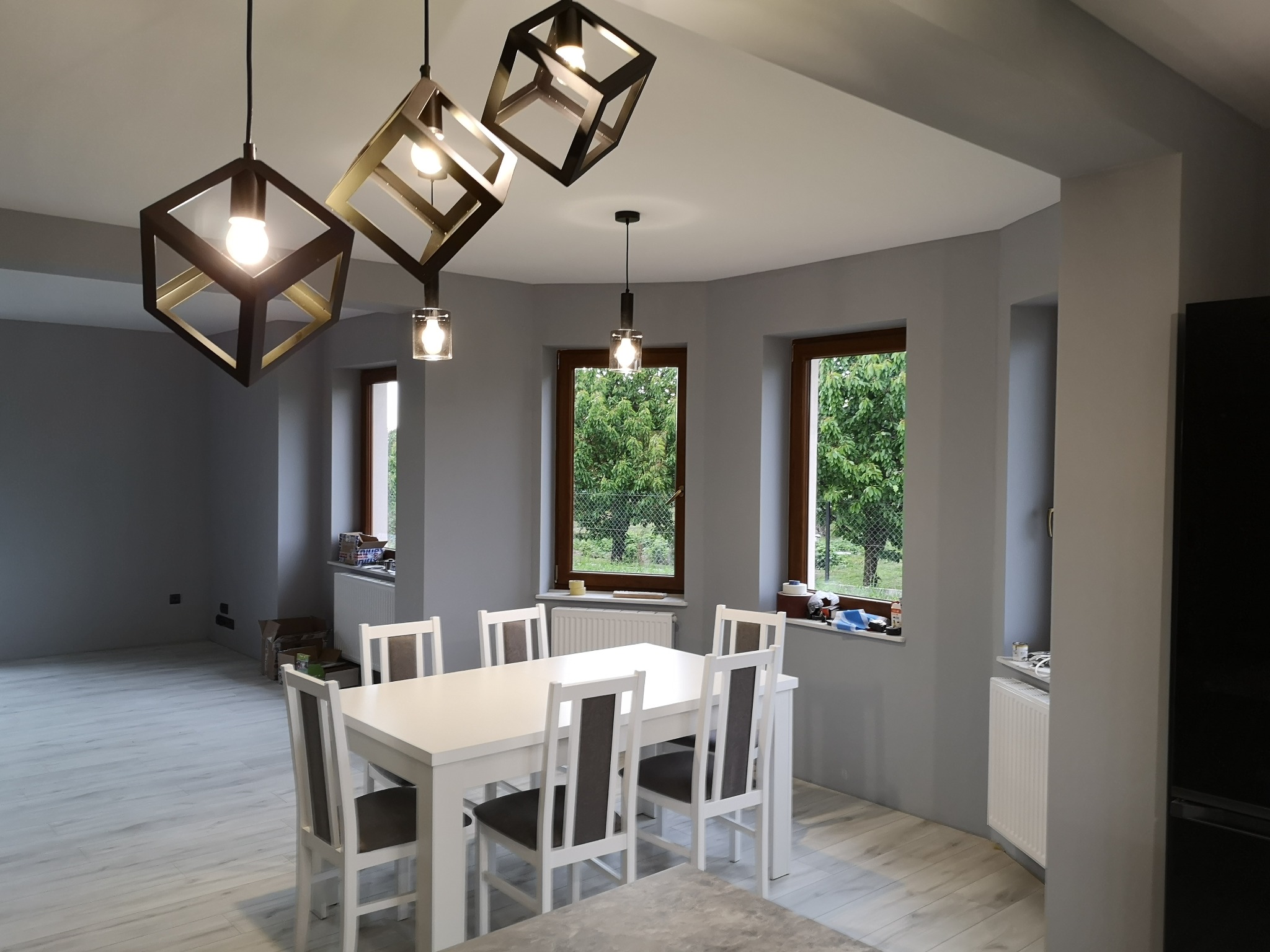 Jasnoszara jadalnia z białym stołem i krzesłami, drewnianymi oknami z widokiem na zieleń oraz trzema nowoczesnymi lampami w kształcie sześcianów.