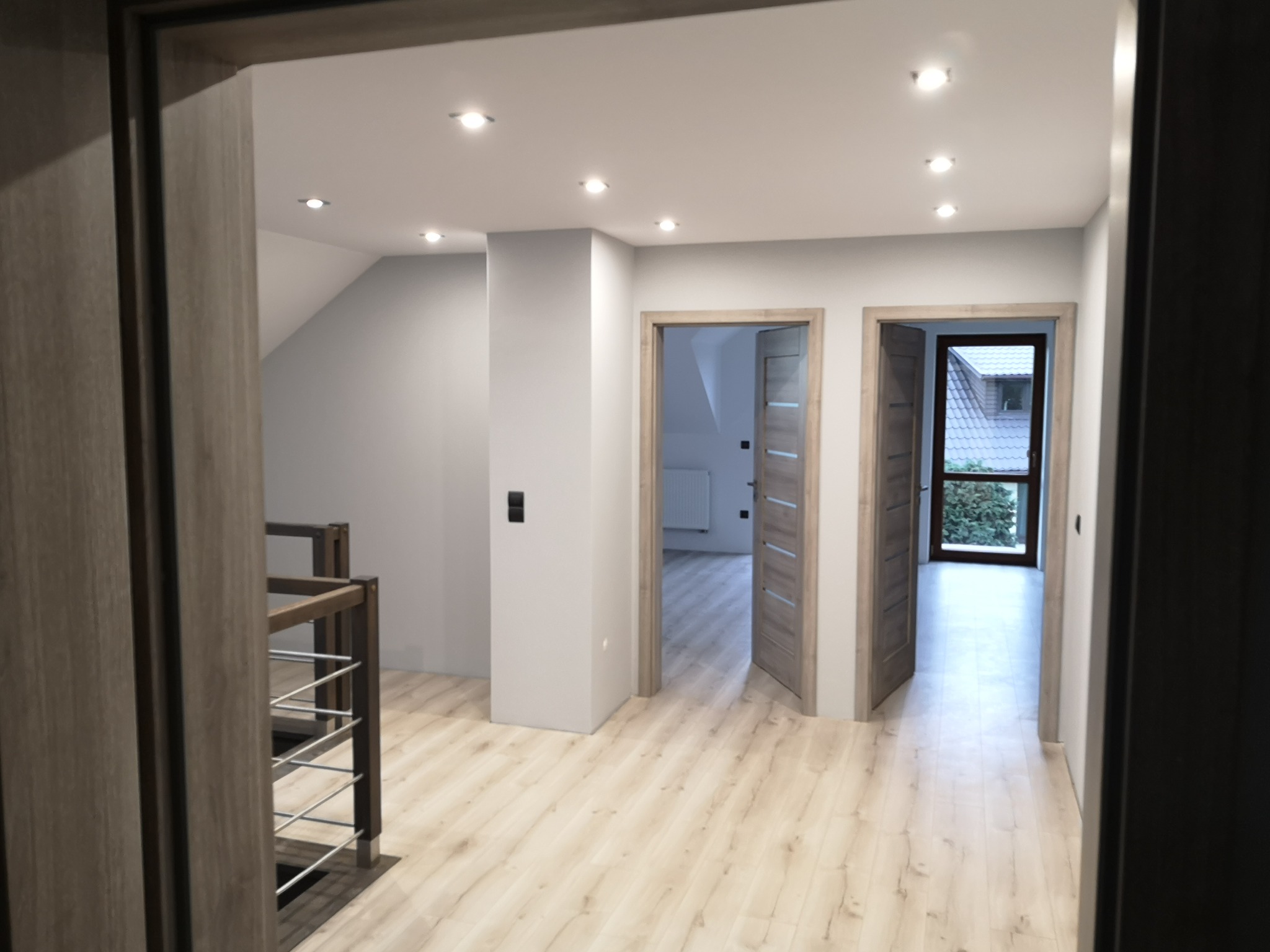 Jasne, przestronne wnętrze z drewnianą podłogą, widok na dwa otwarte pokoje przez drewniane ościeżnice, białe ściany i sufit z punktowym oświetleniem, fragment balustrady po lewej stronie.