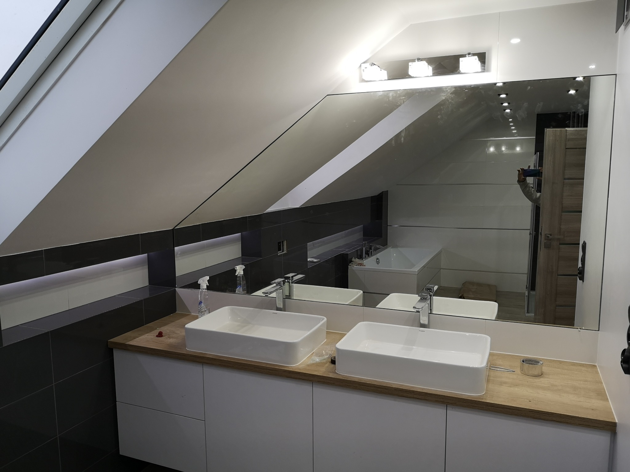 Nowoczesna łazienka na poddaszu z dwoma białymi umywalkami nablatowymi, dużym lustrem, szarą glazurą i oświetleniem punktowym.