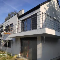 Nowoczesny dom z balkonem zabezpieczonym balustradą ze stali nierdzewnej; widoczne rusztowania sugerują prace wykończeniowe.
