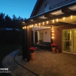 Oświetlony taras z pergolą, wieczorny klimat podkreślony przez wiszące żarówki, widoczne meble ogrodowe i donice z czerwonymi kwiatami, Bolszewo.