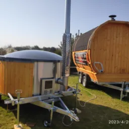 Mobilne jacuzzi z piecem na drewno i sauna w kształcie beczki na przyczepie, ustawione na trawniku w słoneczny dzień.