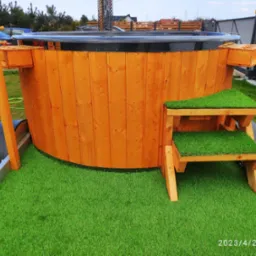 Drewniana balia z hydromasażem i piecem na drewno, umieszczona na przyczepie z zieloną sztuczną trawą, schodki również pokryte sztuczną trawą, widoczne elementy konstrukcyjne i uchwyty po bokach.