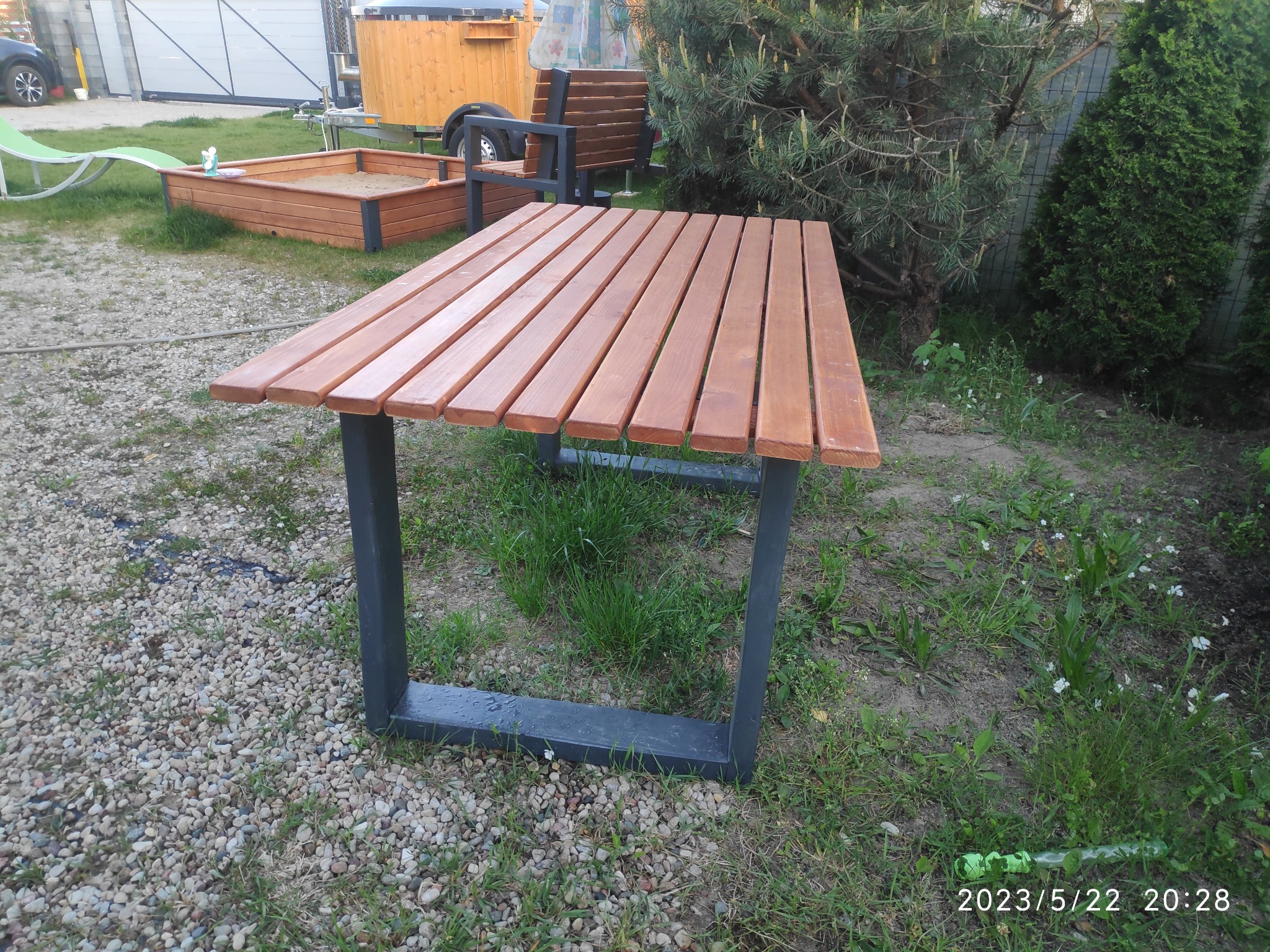 Stół ogrodowy z drewnianym blatem z desek i ciemnymi metalowymi nogami, ustawiony na żwirowym podłożu w ogrodzie, w tle piaskownica i drewniana ławka.