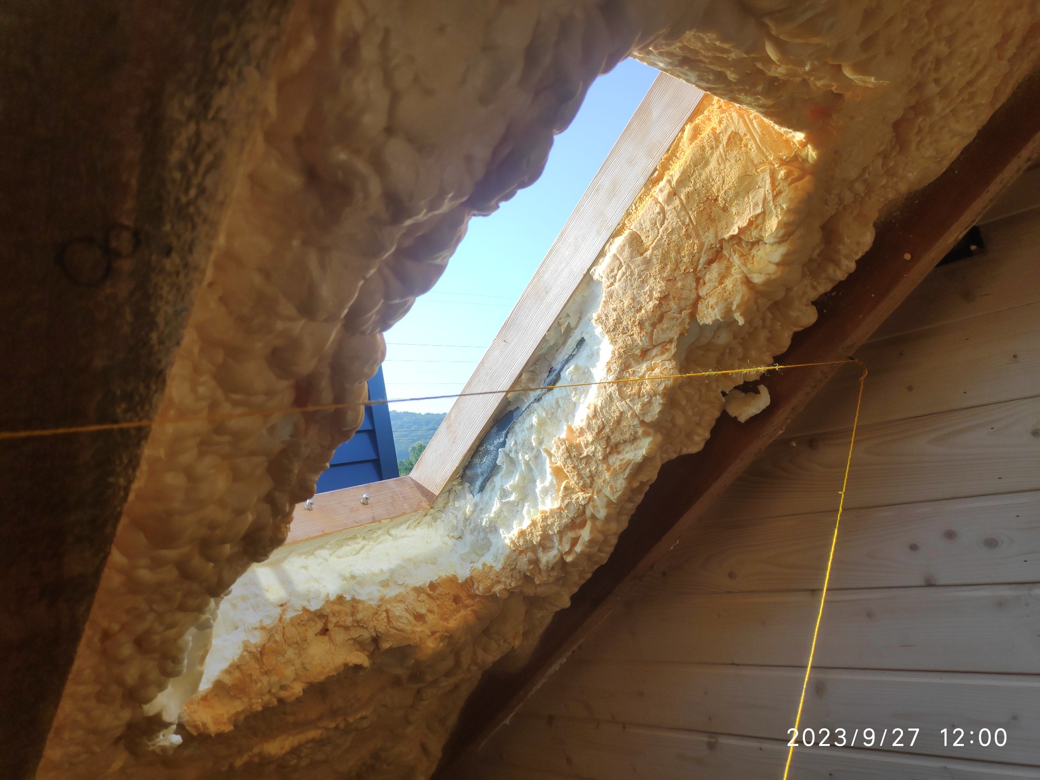 Poddasze w trakcie izolacji pianą PUR, widok z wnętrza przez otwór okienny na błękitne niebo i zielone wzgórza w oddali, widoczne drewniane elementy konstrukcyjne i żółty sznurek wyznaczający linię.