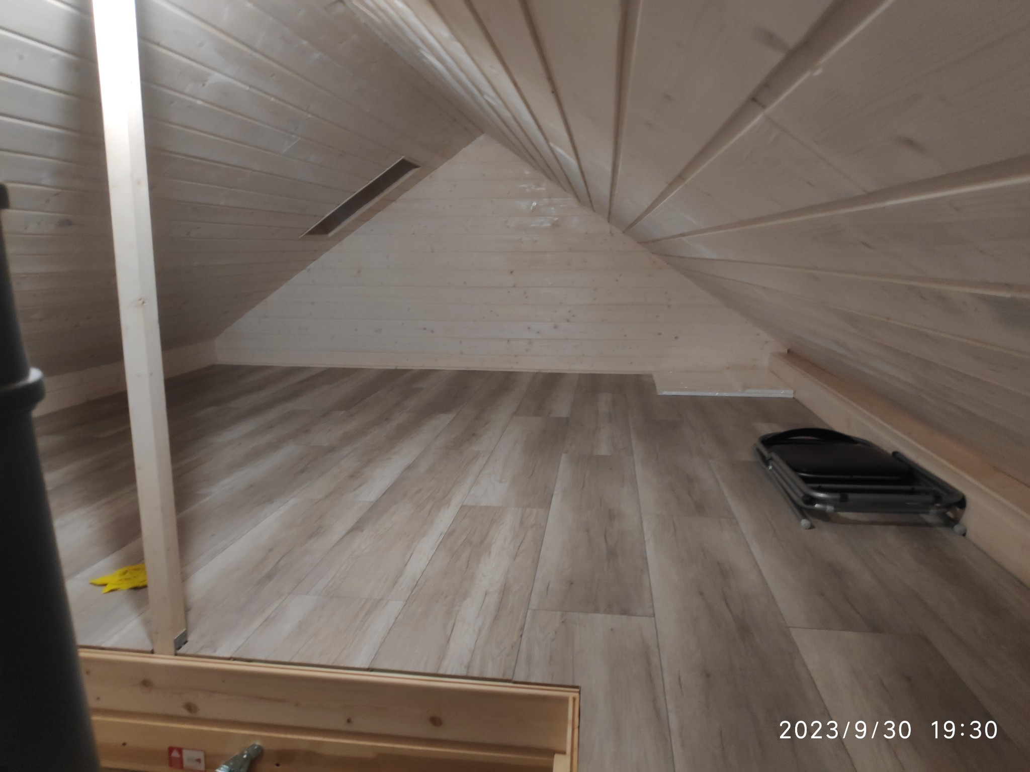 Wykończone poddasze w drewnie z widoczną podłogą z paneli imitujących drewno, skośny sufit z drewnianą boazerią, wentylacja i składane krzesło.