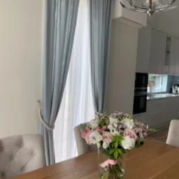 Jasnoszare zasłony z dekoracyjnym sznurem, delikatna firanka, bukiet kwiatów w szklanym wazonie na drewnianym stole, w tle jasna kuchnia i nowoczesny żyrandol.