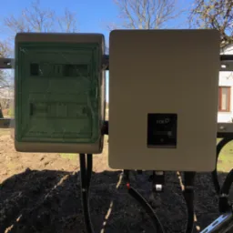 Zewnętrzna instalacja fotowoltaiczna: skrzynka elektryczna w zielonej folii ochronnej i falownik FOX, zamontowane na konstrukcji, z widocznymi przewodami i fragmentem budynku w tle.