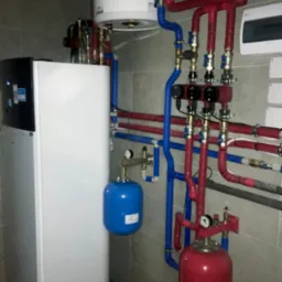 Nowoczesna instalacja grzewcza z białą pompą ciepła, czerwonymi i niebieskimi rurami oraz zbiornikami wyrównawczymi, zamontowana na kafelkowanej ścianie.