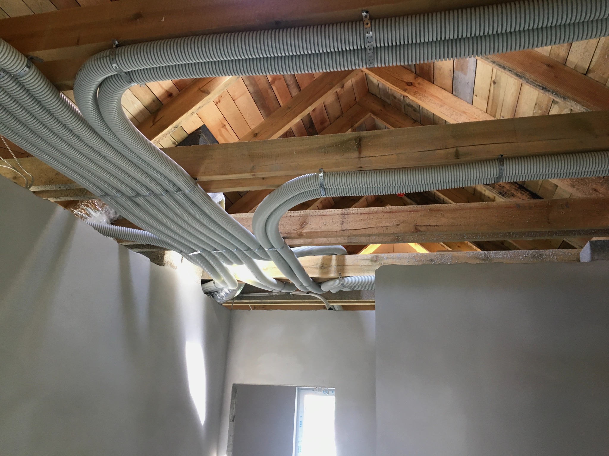 Szare, karbowane rury wentylacyjne zamontowane pod drewnianą konstrukcją dachu w surowym wnętrzu z otynkowanymi ścianami i oknem.