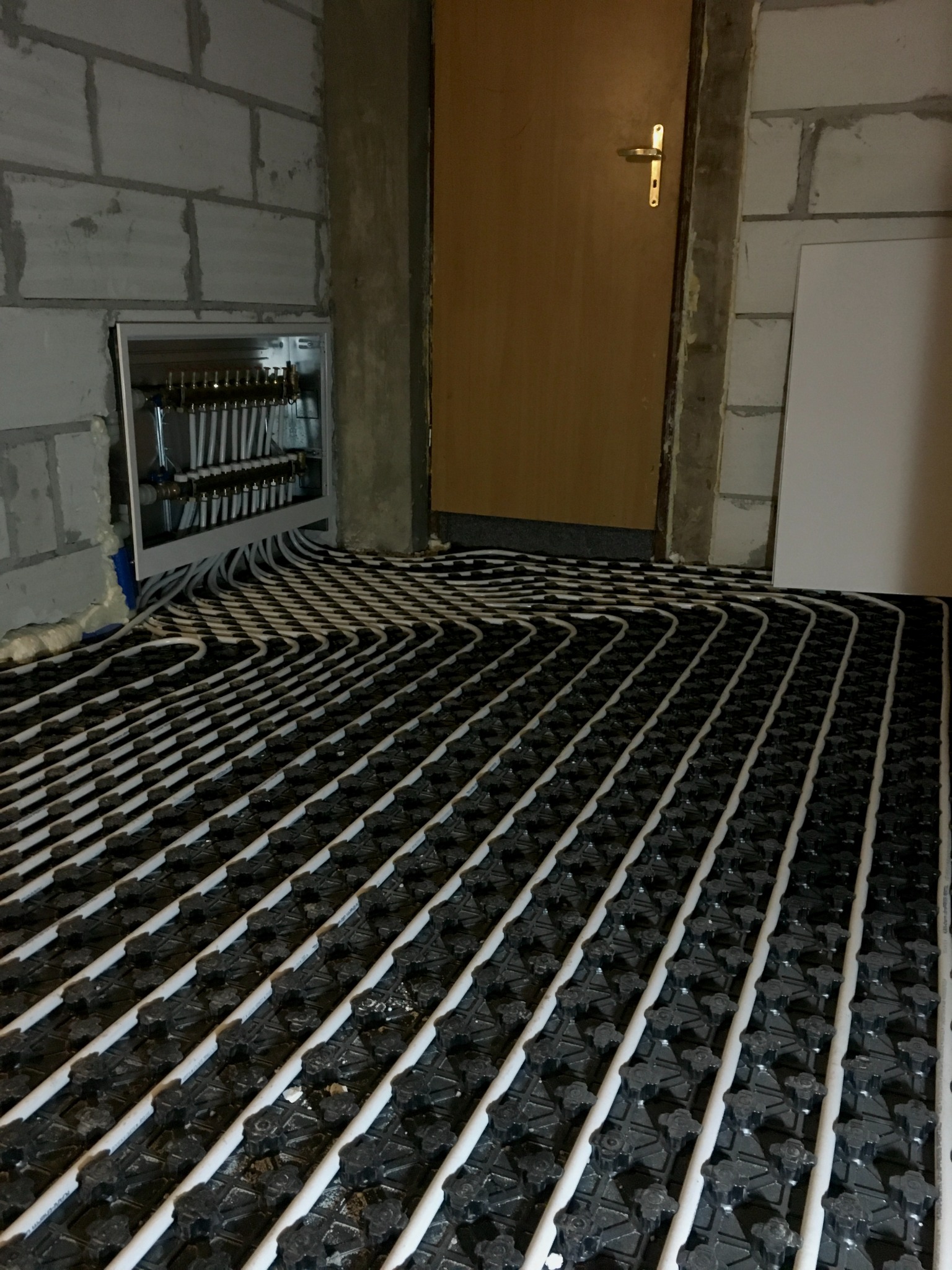 Białe rury grzewcze ułożone w regularnych odstępach na czarnej macie w trakcie realizacji instalacji ogrzewania podłogowego, widoczny rozdzielacz w ścianie z betonu komórkowego i fragment drzwi...