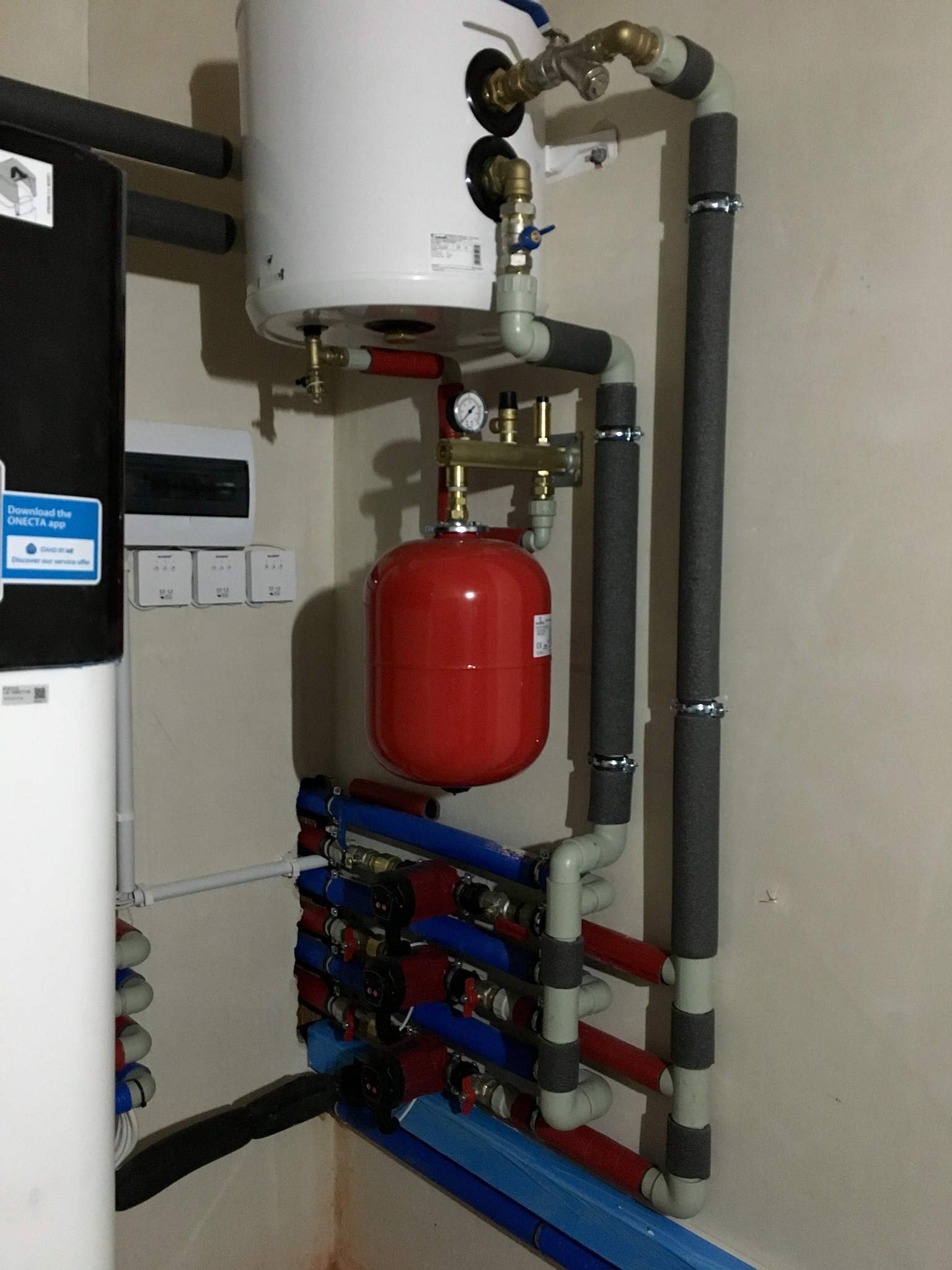 Kocioł z instalacją hydrauliczną, czerwony zbiornik wyrównawczy, rury z izolacją termiczną, rozdzielacz z zaworami i pompami obiegowymi, zamontowany na ścianie w pomieszczeniu.