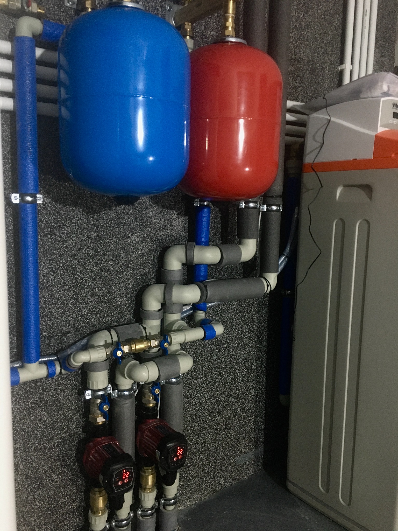 Instalacja grzewcza z dwoma zbiornikami wyrównawczymi (czerwony i niebieski), rurami z izolacją, pompami obiegowymi z wyświetlaczami, zaworami i szarą obudową urządzenia grzewczego.