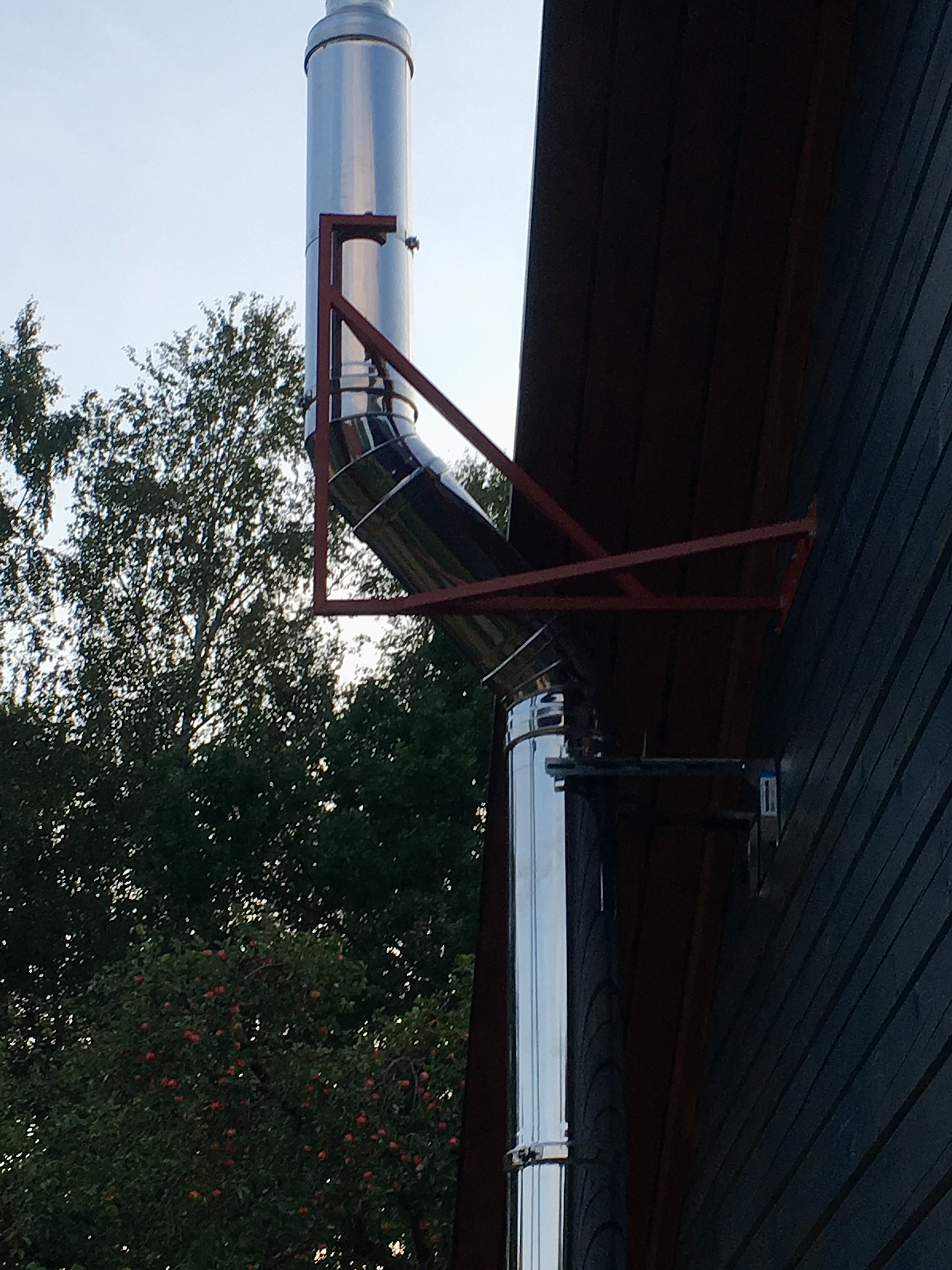 Zewnętrzny, stalowy komin spalinowy zamontowany przy ciemnej, drewnianej ścianie budynku, wsparty na czerwonym, metalowym stelażu.