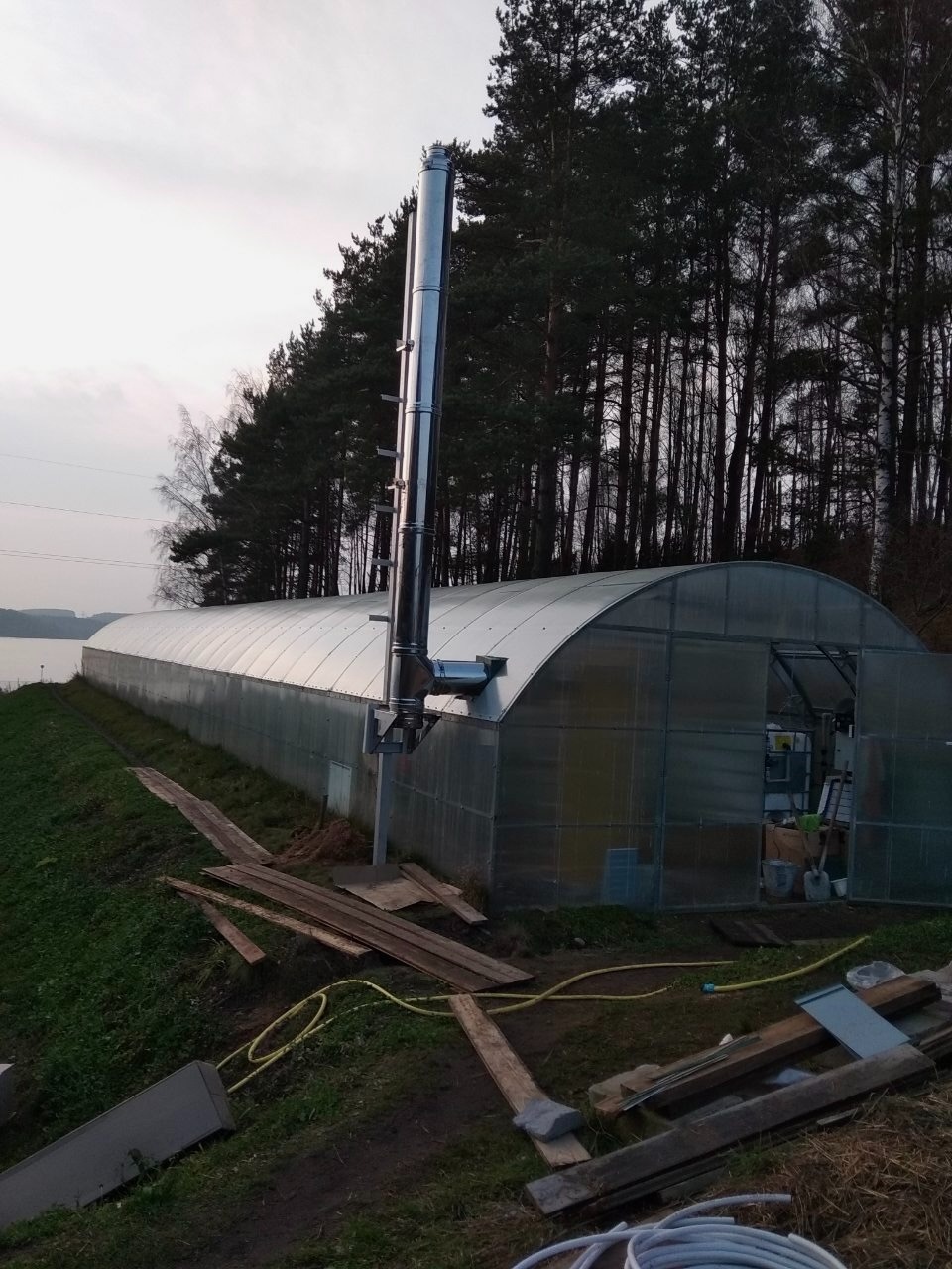 Długi tunel foliowy z kominem spalinowym na zewnątrz, widok z boku, teren nieuporządkowany z deskami i wężami, w tle drzewa i zbiornik wodny.