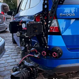 Profesjonalna kamera zamontowana na specjalnym stelażu przymocowanym do tylnej klapy niebieskiej Skody Fabii, widok z bliska na sprzęt i fragment samochodu na tle brukowanej ulicy.