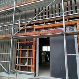 Budynek w trakcie budowy z widoczną drewnianą konstrukcją szkieletową, deskowaniem zewnętrznym i rusztowaniem z logo firmy na belkach.